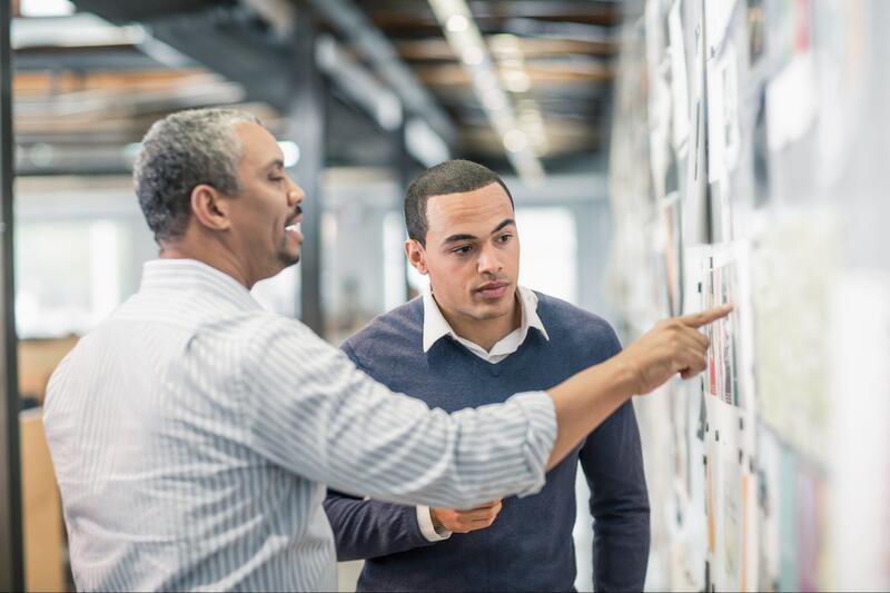 Two men talking and one of them pointing at a board